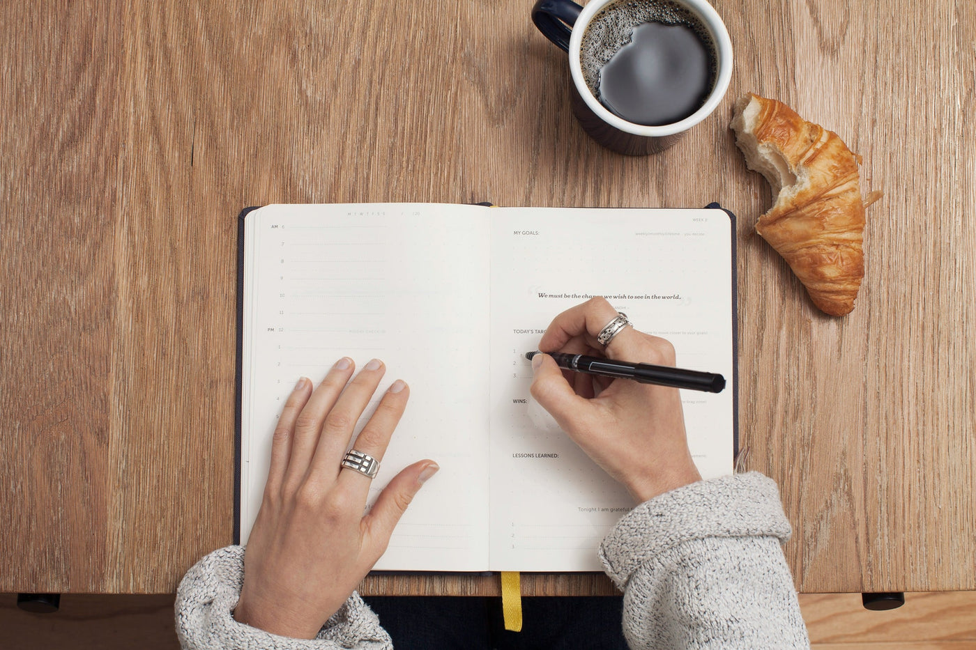 Image shows hands writing in a journal with a coffee and croissant next to them.